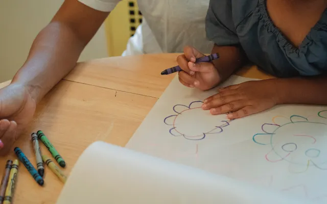 Criança desenhando à mesa com o apoio de uma pessoa adulta.